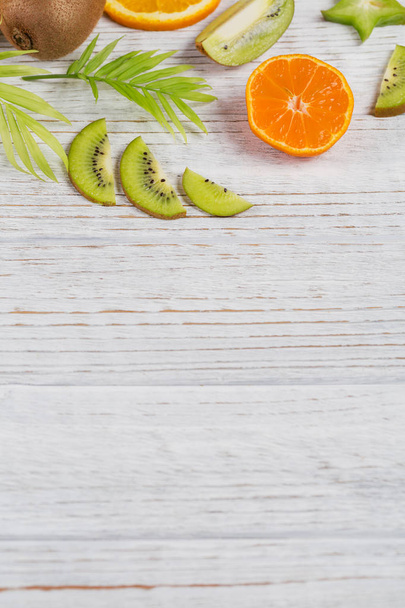 Green palm leaves and fresh tropical slices of fruits on white wooden table. Juicy oranges, tangerines, limes, kiwis, carambolas. Summer fruit background. Flat lay, top view, copy space - 写真・画像