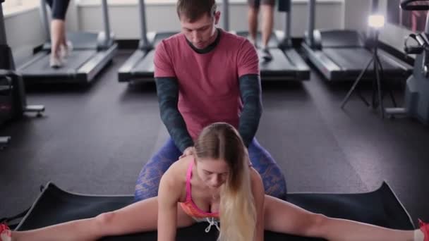 Athletic and attractive man helps a beautiful woman to stretch on the floor in the gym. - Footage, Video
