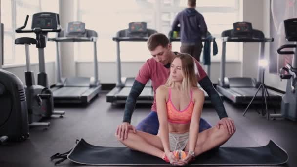athletischer und attraktiver Mann hilft einer schönen Frau, sich im Fitnessstudio auf dem Boden zu strecken. - Filmmaterial, Video