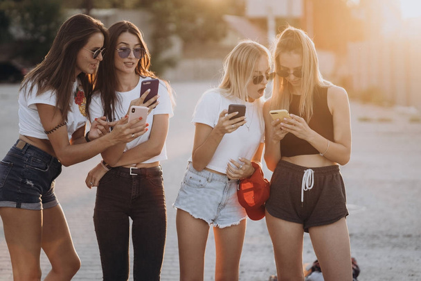 Four attractive women are standing on car parking with smartphones - Фото, изображение