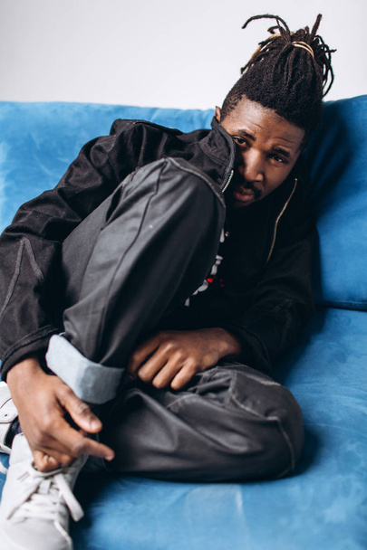 One attractive smiling black man in a black jacket and black pants on a gray background sidin on a blue sofa - Foto, afbeelding