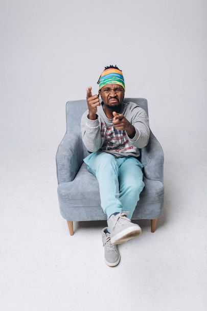 Portrait of a smiling black young model in a gray sweater and blue pants on a gray background near the chair - Foto, Imagem