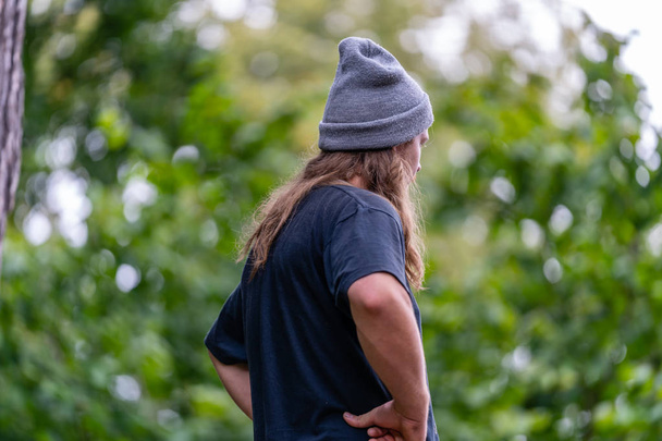 Foto des Hippie-Millennials mit blauem T-Shirt, grauem Hut und langen Haaren, der seine Freizeit im Park auf dem Land verbringt - Konzept des gebildeten Generationenmitglieds, freier Raum für Text - Foto, Bild