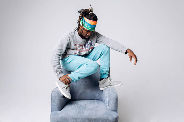 Portrait of a smiling black young model in a gray sweater and blue pants on a gray background near the chair - Foto, imagen
