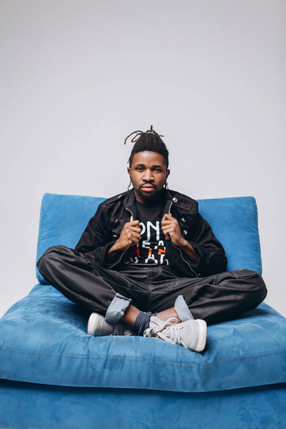 One attractive smiling black man in a black jacket and black pants on a gray background sidin on a blue sofa - Fotografie, Obrázek