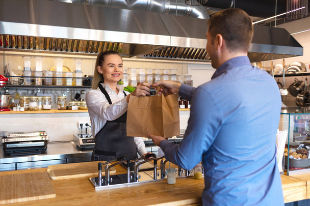 Small business and entrepreneur concept with smiling young waitress wearing black apron serving customer at counter in family restaurant, takeaway food. Small family business  - Φωτογραφία, εικόνα
