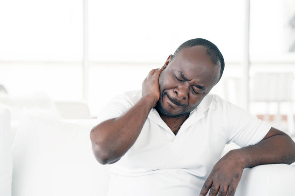 Tired neck. Man suffering from neck pain at home on couch. Males sense of fatigue, exhausted, stressed. African man massages her painful neck with her hands. The concept of body and health. - Fotoğraf, Görsel