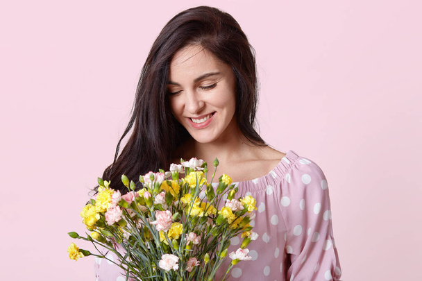 Positive of young European woman with dark hair, holds pretty bouquet, smiles gently, wears polka dot fashionable clothes, prepares for International Womens Day, isolated over pink background - Фото, зображення