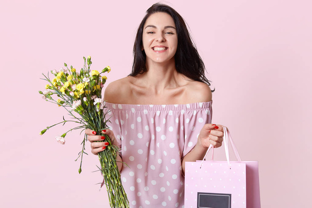 Photo of pleased brunette European lady holds gift bag and flowers, happy to recieve present on Womens Day, dressed in polka dot dress, shows bare shoulders, has positive look, poses indoor. - Фото, изображение