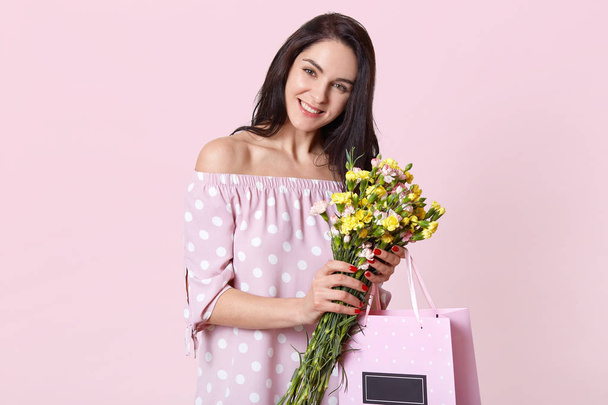 Satisfied good looking woman with dark hair, smiles broadly, holds pretty flowers and gift bag wears summer dress, looks directly at camera with happy expression, models over light pink background - Фото, изображение