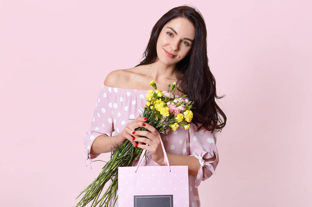 Isolated shot of attractive young European woman has black long hair, wears polka dot dress, holds gift bag and flowers, poses over light pink studio wall, celebrates International Womens day - Fotografie, Obrázek