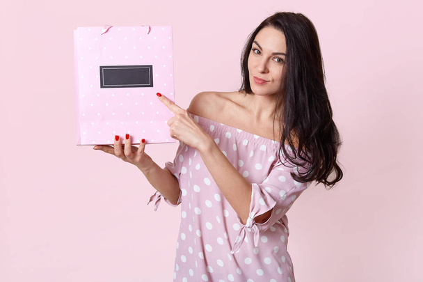 Horizontal view of pleasant looking European woman points at gift bag, shows free space for your advertising content or promotion, dressed in polka dot dress, has red manicure. Isolated shot - Φωτογραφία, εικόνα