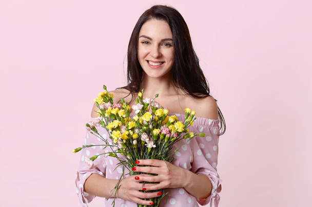 Happy positive woman with dark hair, holds flowers in hands, smiles positively, enjoys spring warm day, dressed in stylish polka dot dress, isolated over pink background. Ladies like flowers - Zdjęcie, obraz