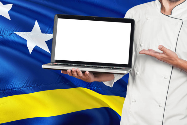 Chef holding laptop with blank screen on Curacao flag background. Cook wearing uniform and pointing laptop for copy space. - Fotó, kép