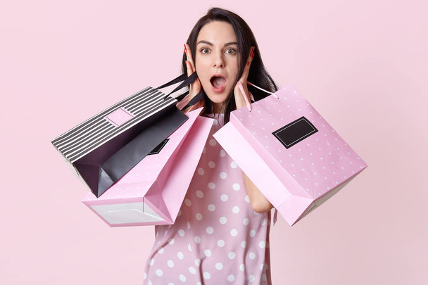 Image of attractive shocked woman with dark hair, carries shopping bags, keeps hands on cheeks, dressed in polka dot dress, surprised with big discounts in shopping mall, poses on pink wall. - Photo, image