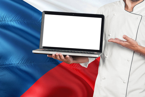 Czech Chef holding laptop with blank screen on Czech Republic flag background. Cook wearing uniform and pointing laptop for copy space. - Fotó, kép