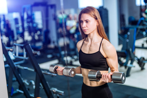 Sporty woman doing exercises with lifting dumbbells looking ahead on gym background. Photo of muscular female wearing sportswear working out. Strength and motivation - Foto, Bild