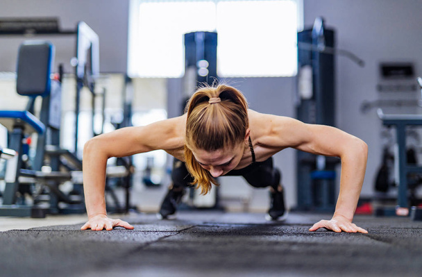 Beautiful girl does push-ups from the floor for training the muscles of the hands in the gym on the background of sports equipment. Sportswoman with hair in ponytail is doing muscle training - Photo, image