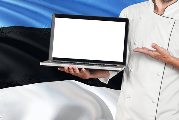 Estonian Chef holding laptop with blank screen on Estonia flag background. Cook wearing uniform and pointing laptop for copy space. - Fotó, kép