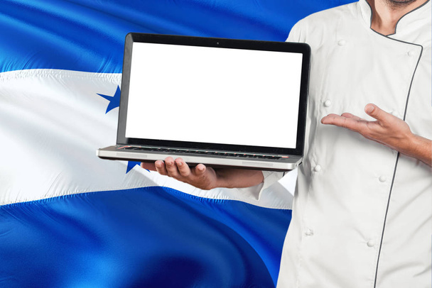 Honduran Chef holding laptop with blank screen on Honduras flag background. Cook wearing uniform and pointing laptop for copy space. - Fotó, kép