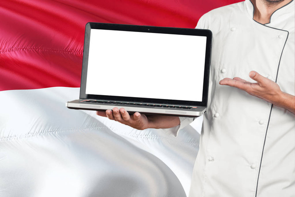Indonesian Chef holding laptop with blank screen on Indonesia flag background. Cook wearing uniform and pointing laptop for copy space. - Fotó, kép