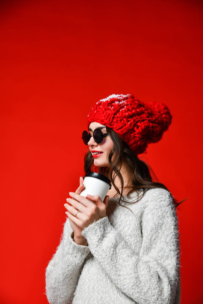 Close up portrait of a smiling young girl in hat holding take away coffee cup over red wall - Фото, изображение
