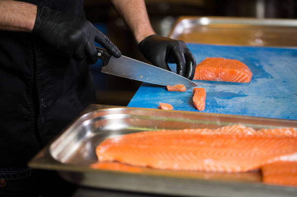 テーマの料理は、調理の専門職です。赤い魚を準備するレストランのキッチンでの白人男性の手のクローズ アップ黒ラテックス手袋制服でサーモン肉を切り身. - 写真・画像
