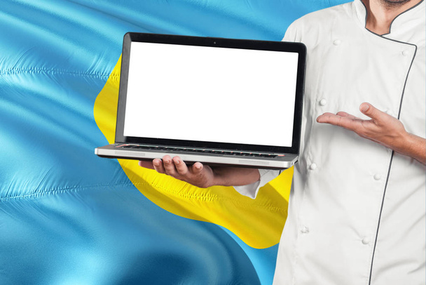 Palauan Chef holding laptop with blank screen on Palau flag background. Cook wearing uniform and pointing laptop for copy space. - Fotó, kép