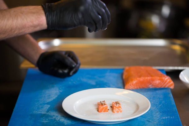 テーマの料理は、調理の専門職です。赤い魚を準備するレストランのキッチンでの白人男性の手のクローズ アップ黒ラテックス手袋制服でサーモン肉を切り身. - 写真・画像