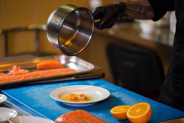Θέμα το μαγείρεμα είναι ένα επάγγελμα του μαγειρέματος. Κοντινό πλάνο του χέρι ενός άνδρα Καυκάσιος σε κουζίνα εστιατορίου προετοιμασία κόκκινα ψάρια φιλέτα σολομού κρέας σε μαύρα γάντια λατέξ, στολή. - Φωτογραφία, εικόνα