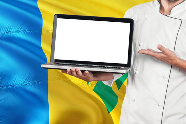 Chef holding laptop with blank screen on Saint Vincent And The Grenadines flag background. Cook wearing uniform and pointing laptop for copy space. - Fotó, kép