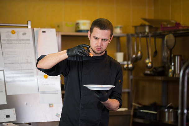 Θέμα το μαγείρεμα είναι επάγγελμα του μαγειρέματος. Πορτρέτο του Καυκάσου άνθρωπος σε κουζίνα εστιατορίου προετοιμασία κόκκινα ψάρια φιλέτα σολομού κρέας σε ομοιόμορφο μαύρο γάντια. Σεφ κατέχει πλάκα με μπλε και ελέγχους, γεύσεις. - Φωτογραφία, εικόνα
