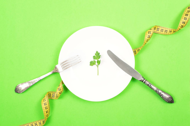 Diet, weigh loss, healthy eating, fitness concept. Small portion of food on big plate. Small green salad leaf on white plate with fork and knife on the background of measuring tape. - Photo, Image
