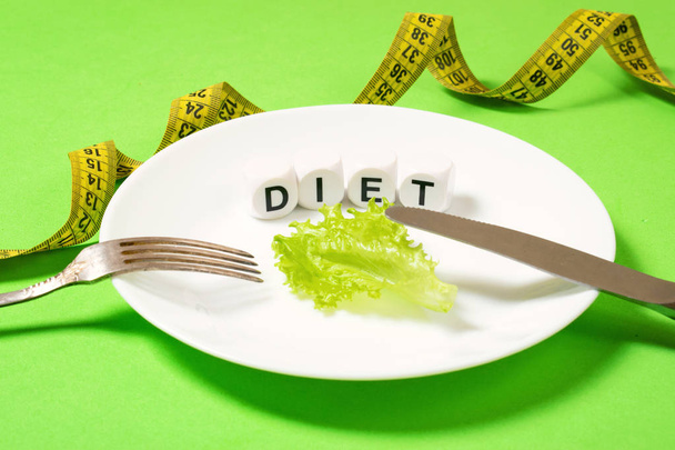 Diet, weigh loss, healthy eating, fitness concept. Small portion of food on big plate. Small green salad leaf on white plate with fork and knife and text diet on the background of measuring tape. - Photo, Image