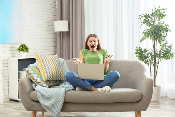 Emotional young woman with laptop celebrating victory on sofa at home - Φωτογραφία, εικόνα