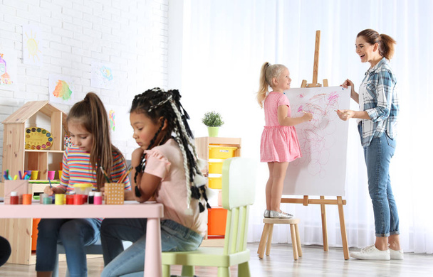 Учительница с ребенком возле мольберта на уроке живописи в помещении
 - Фото, изображение