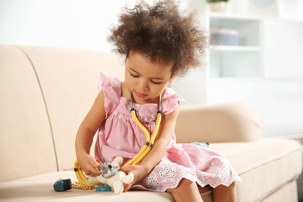 Schattig African American kind verbeelden zich als arts tijdens het spelen met de stethoscoop en doll op Bank thuis - Foto, afbeelding