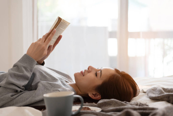 Smiling Beautiful Asian woman reading a book while lying on a bed. Vacation or Holiday. - 写真・画像