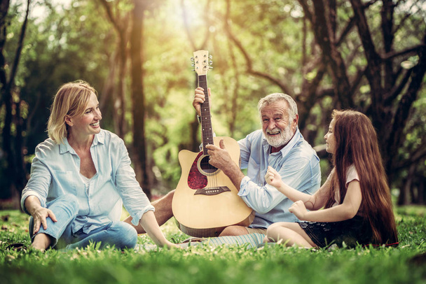 Happy family play guitar and sing together while sitting in the park in summer. Concept of family bonding by music. - Zdjęcie, obraz