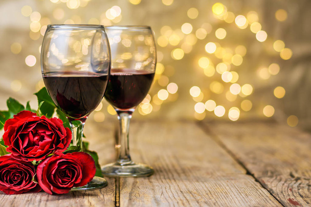 Valentine's Day still life. Two glasses of wine and red roses on a rustic wooden table in front of a blurred luminous background with copy space for text. Selective focus. - 写真・画像