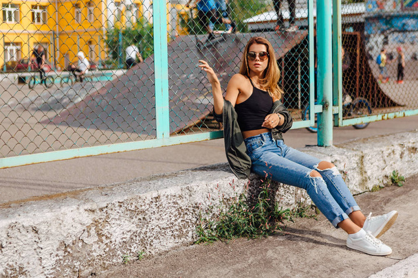 Fashion portrait of trendy young woman wearing sunglasses, jeans with halls and bomber jacket in the city. Copy space. - 写真・画像