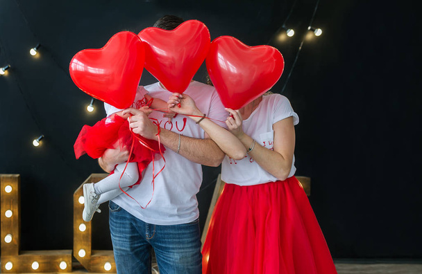 Love and Valentines day. Caucasian family hiding faces behind red heart shaped balloons, black studio background. Horizontal family portrait - Fotografie, Obrázek