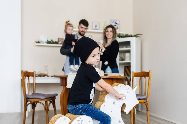 Selectieve aandacht van kleine jongen in een zwarte pet op houten paard speelgoed en familie op een achtergrond. Horizontale portret - Foto, afbeelding