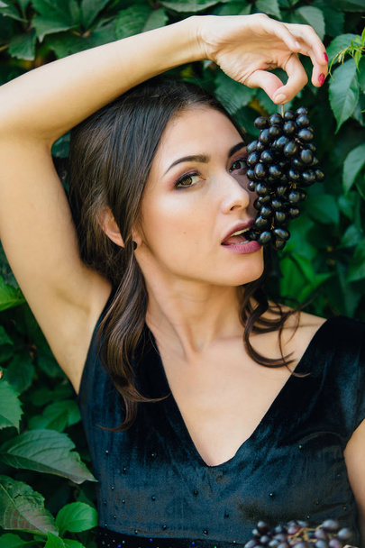 Portrait of young sexy brunette biting black grapes. - Photo, Image