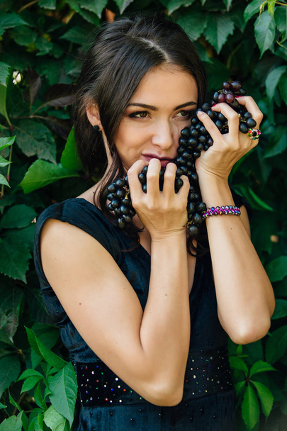 Portrait of young sexy brunette biting black grapes. - Foto, afbeelding