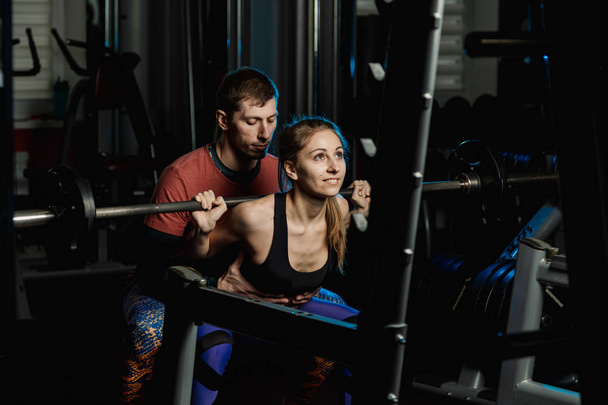 Handsome athletic man helps a beautiful girl to squat with a barbell on the shoulders in the gym. - Photo, Image