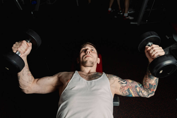 schöner athletischer Mann trainiert Brustmuskeln, indem er Hanteln züchtet, während er im Fitnessstudio liegt. - Foto, Bild