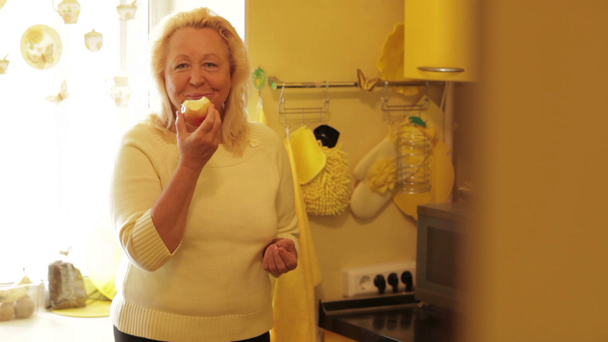 Mujer mayor comiendo manzana en la cocina amarilla y mirando a la cámara
. - Imágenes, Vídeo