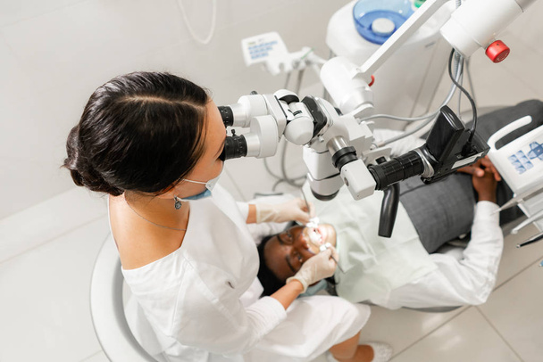 Fiatal nő fogorvos kezelésére gyökércsatornákat használ Mikroszkóp a fogászati klinikán. Férfi beteg feküdt a fogorvos szék, nyitott szájjal. Orvosi, fogorvosi és egészségügyi koncepció. Fogászati berendezés - Fotó, kép