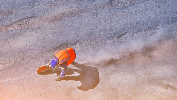 Aerial view of unrecognizable road builder worker cutting asphalt or concrete with saw blade at construction site. cut-off machine power tool breaking asphalt. Lots of dust from cutting. - Фото, изображение